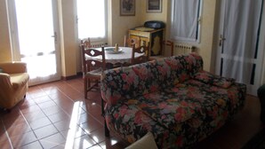 A living room with a floral sofa, a wooden chair, a small table with a plate and a bottle, and a radiator.