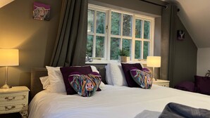 Quarto com uma cama grande, duas almofadas com design colorido, mesa de cabeceira com abajur e uma janela com cortinas.