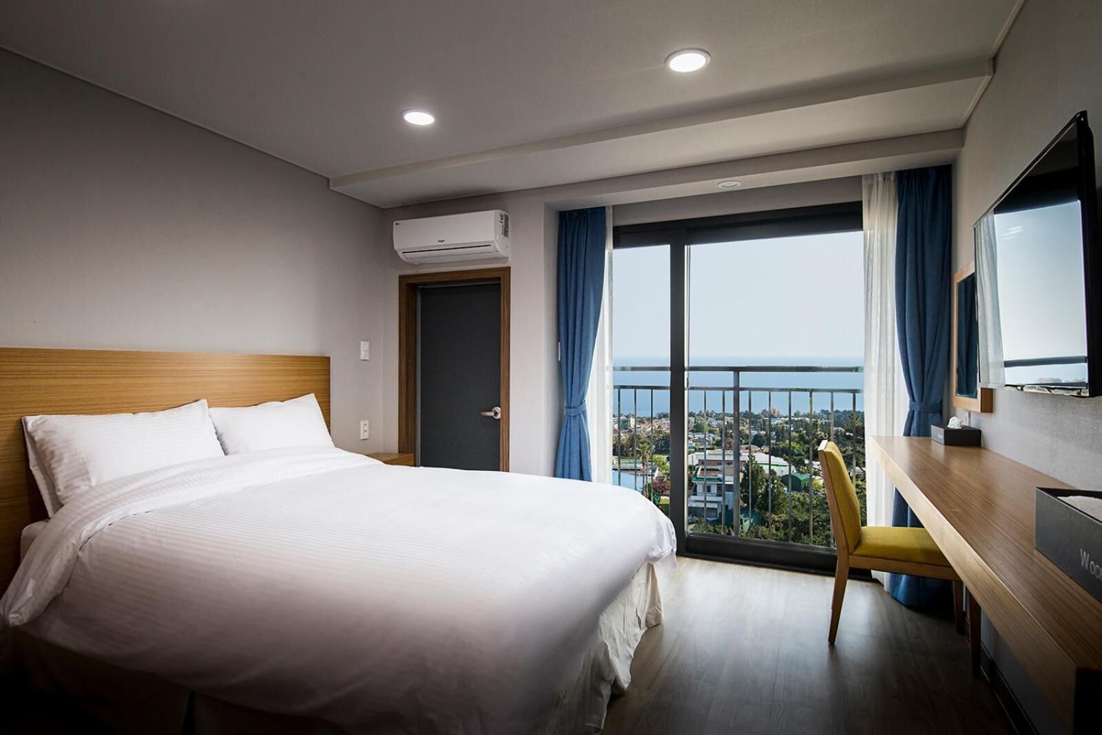 A hotel room with a large bed, a desk, a chair, an air conditioning unit, a television, and a balcony with a view of the city and the sea.