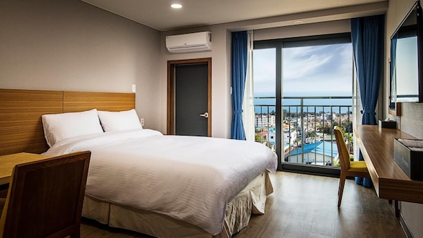 A hotel room with a bed, a desk, and a view of the city through large windows. - W Ocean Pension (Seogwipo)