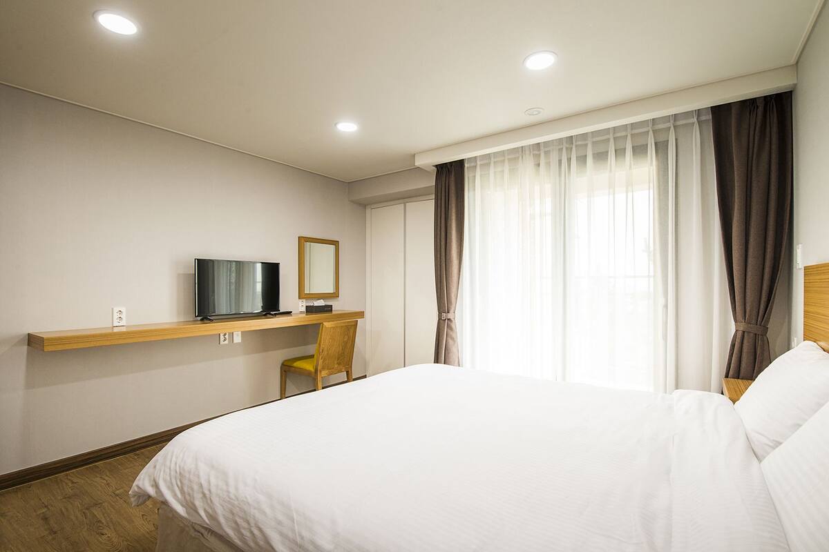 A hotel room with a bed, a desk with a television, a chair, and a window with curtains.