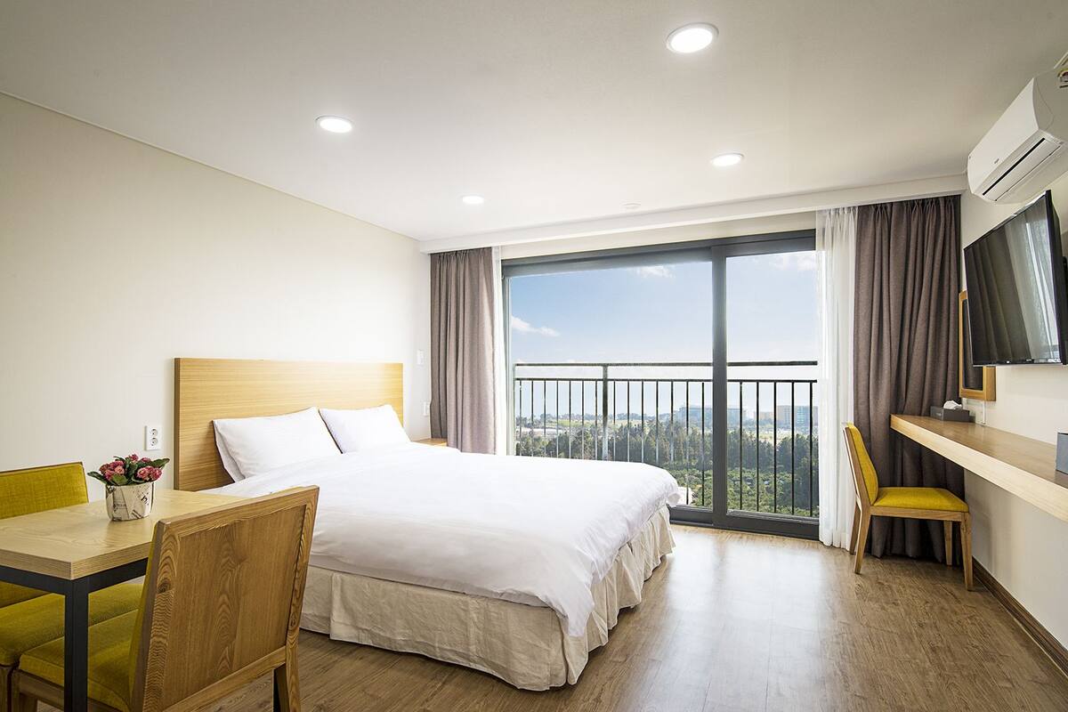 A hotel room with a large bed, a desk, a chair, a TV, and a balcony with a city view.