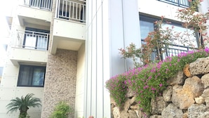 A multi-story building with balconies, a stone wall, and a rock garden with pink flowers. - W Ocean Pension (Seogwipo)