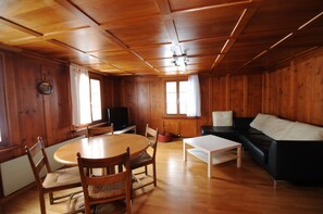 A wooden interior with a round dining table, chairs, a sofa, and a television.
