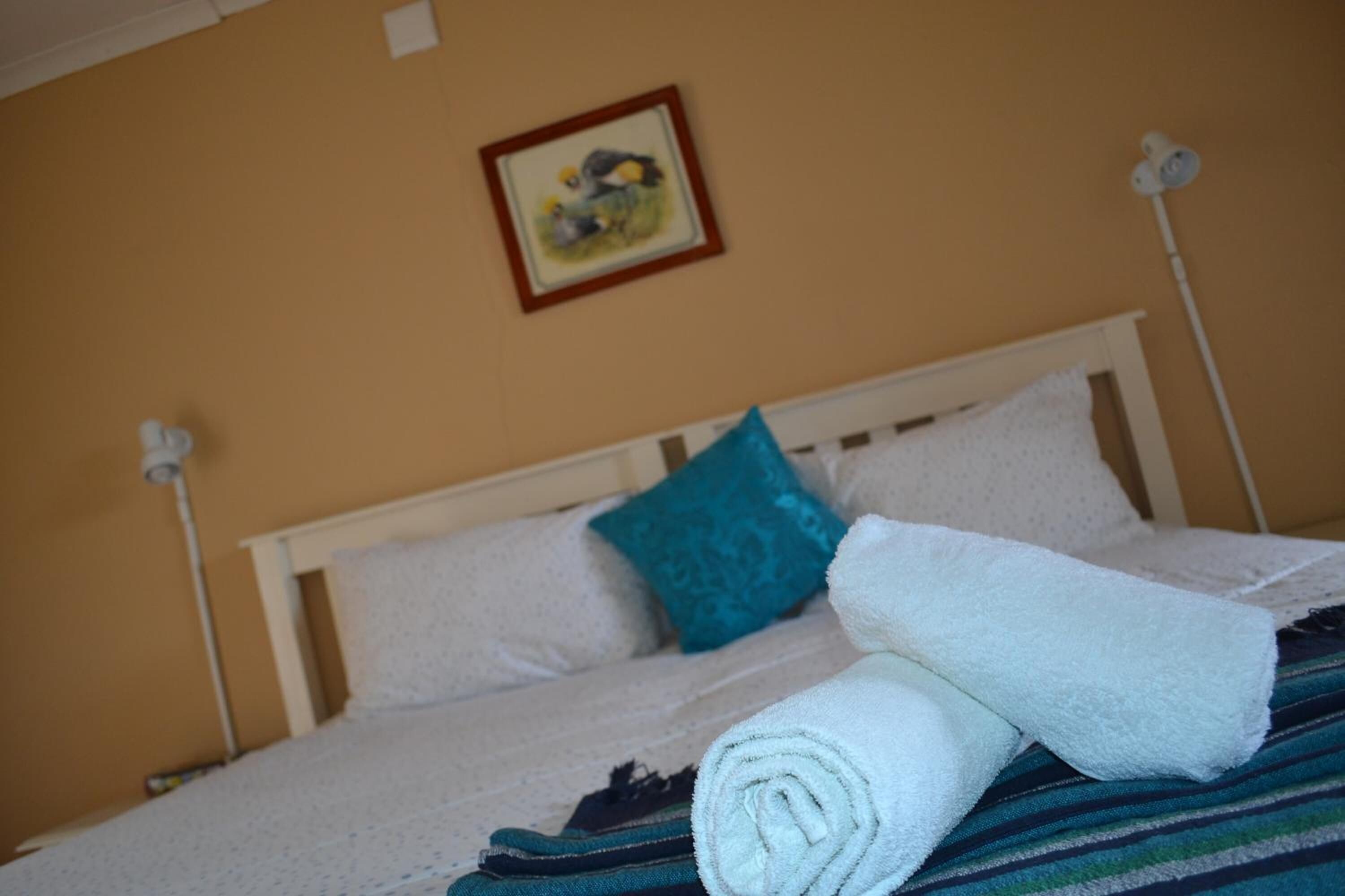 A neatly made bed with white linens, a teal pillow, and a rolled towel on a white headboard.