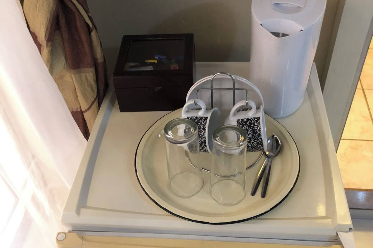 A compact refrigerator with a glass door, a small black box, a white trash bin, and a metal tray with glasses and a spoon.