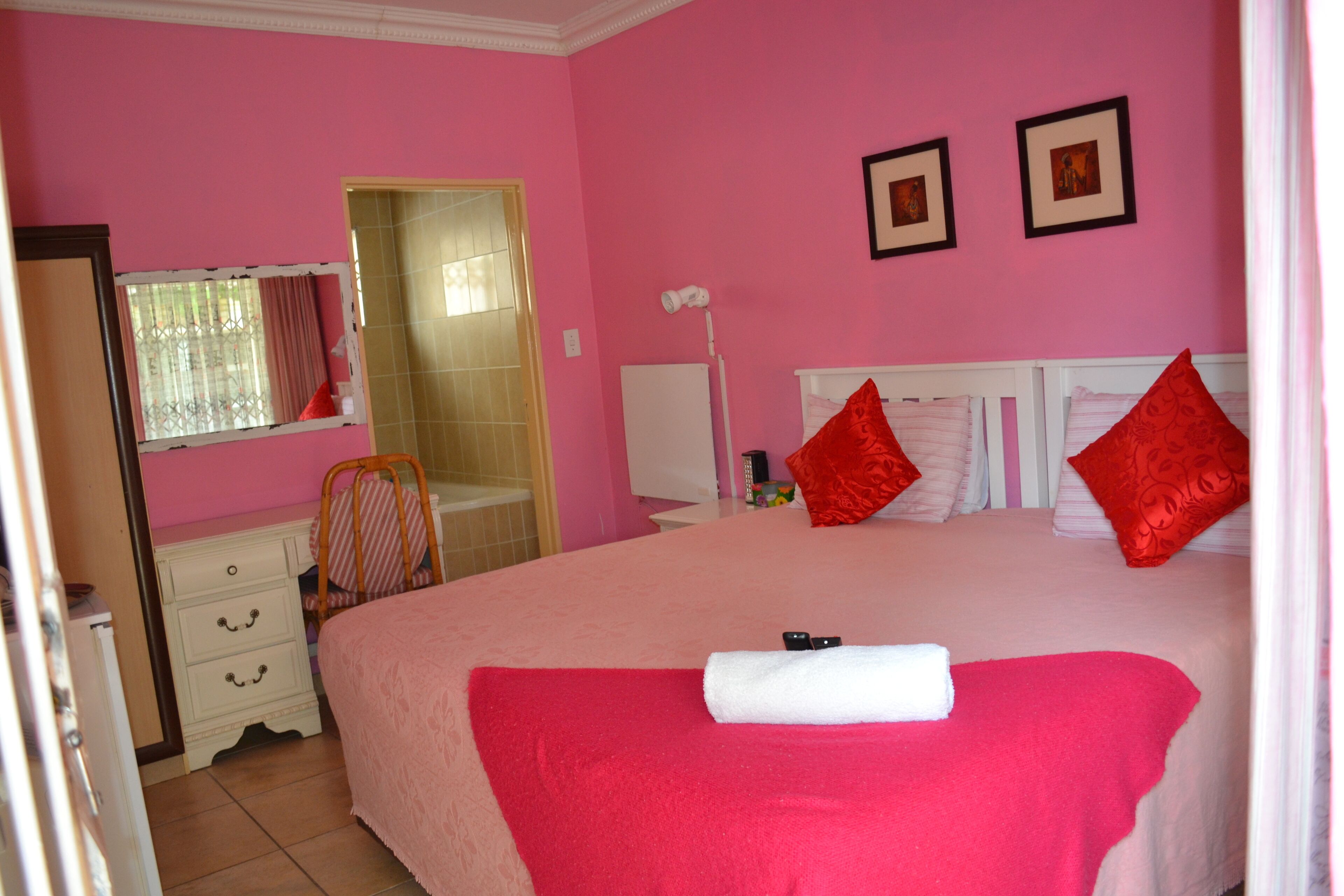 Un dormitorio rosa con una cama grande, almohadas rojas, cabecera blanca y un pequeño escritorio con cajones.