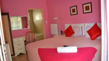 Un dormitorio rosa con una cama grande, almohadas rojas, cabecera blanca y un pequeño escritorio con cajones.
