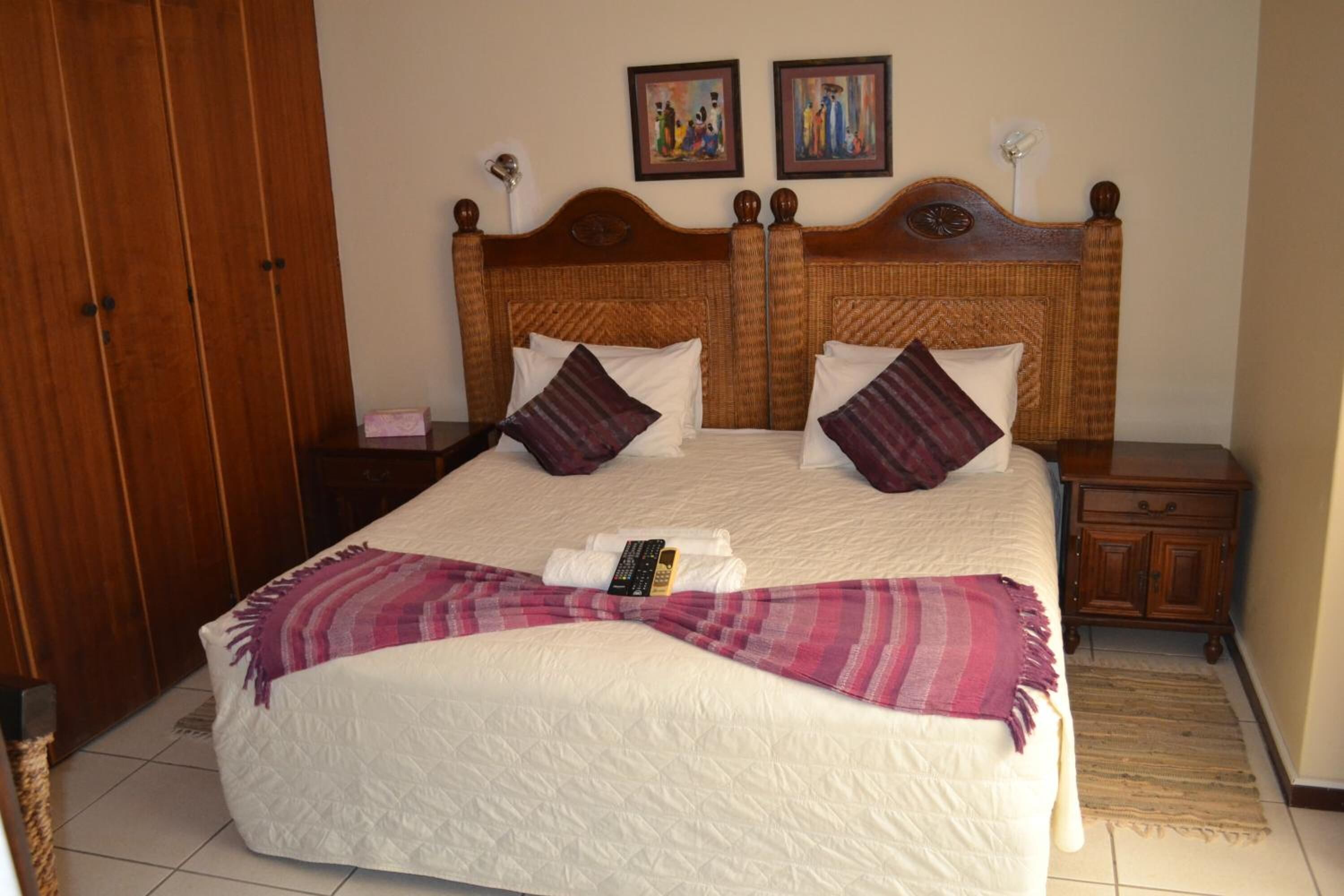 A double bed with a white quilt, two wooden nightstands, a wooden wardrobe, and two paintings on the wall.