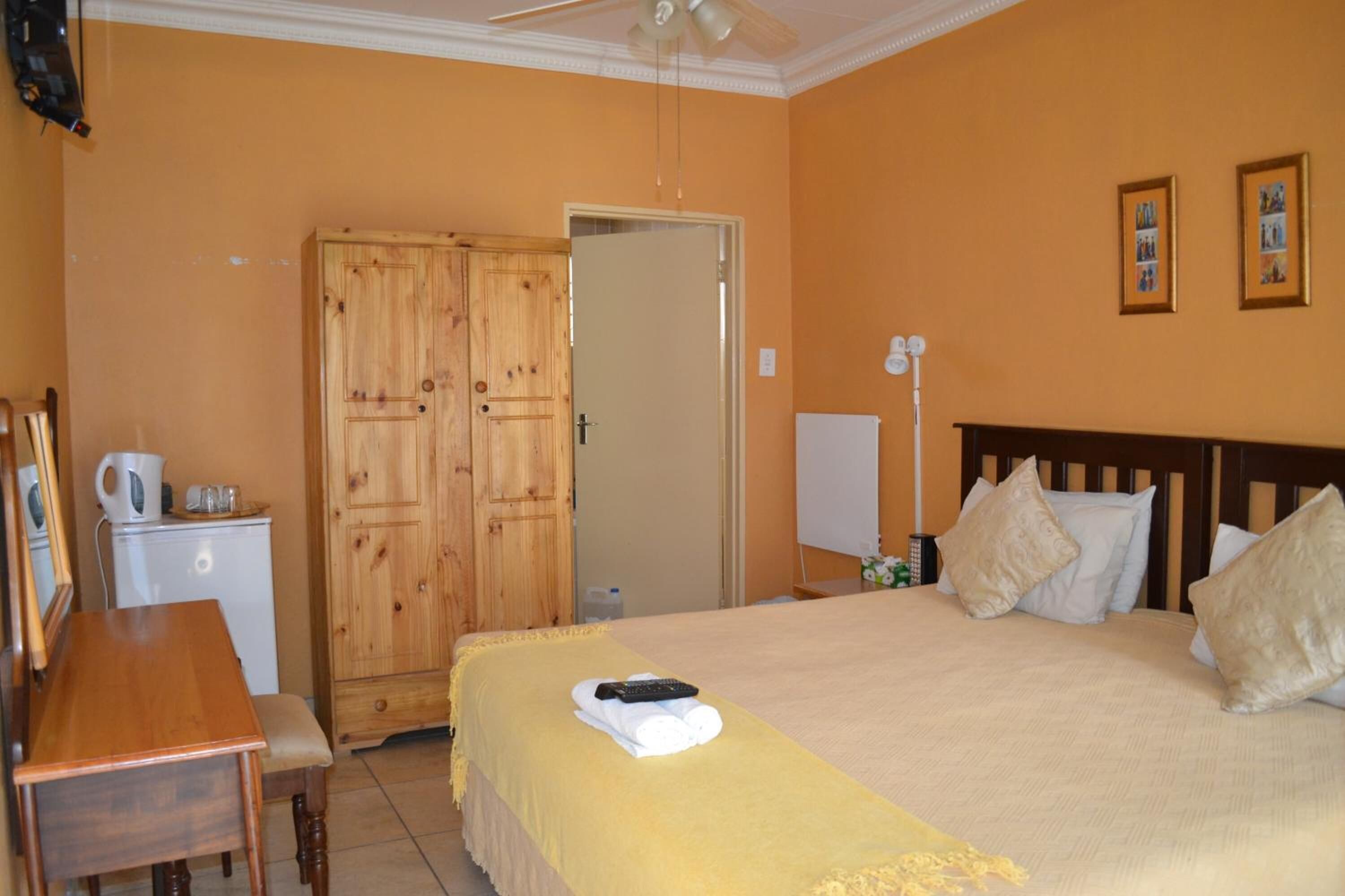 Quarto de hotel com cama, guarda-roupa de madeira, secretária com chaleira e mesa de cabeceira.