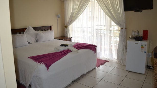 A hotel room with a bed, a television, a refrigerator, and a window with curtains. - Kamogelo Guest House (Moses Kotane)