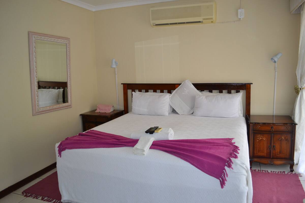 A neatly made bed with white linens and a pink blanket, a bedside table with a lamp, a wooden nightstand, and a mirror on the wall.