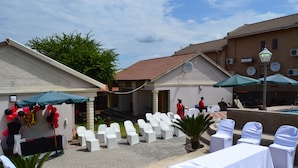 An outdoor event area with white chairs, tables, and umbrellas, a building with a tiled roof, and a clear sky. - Kamogelo Guest House (Moses Kotane)