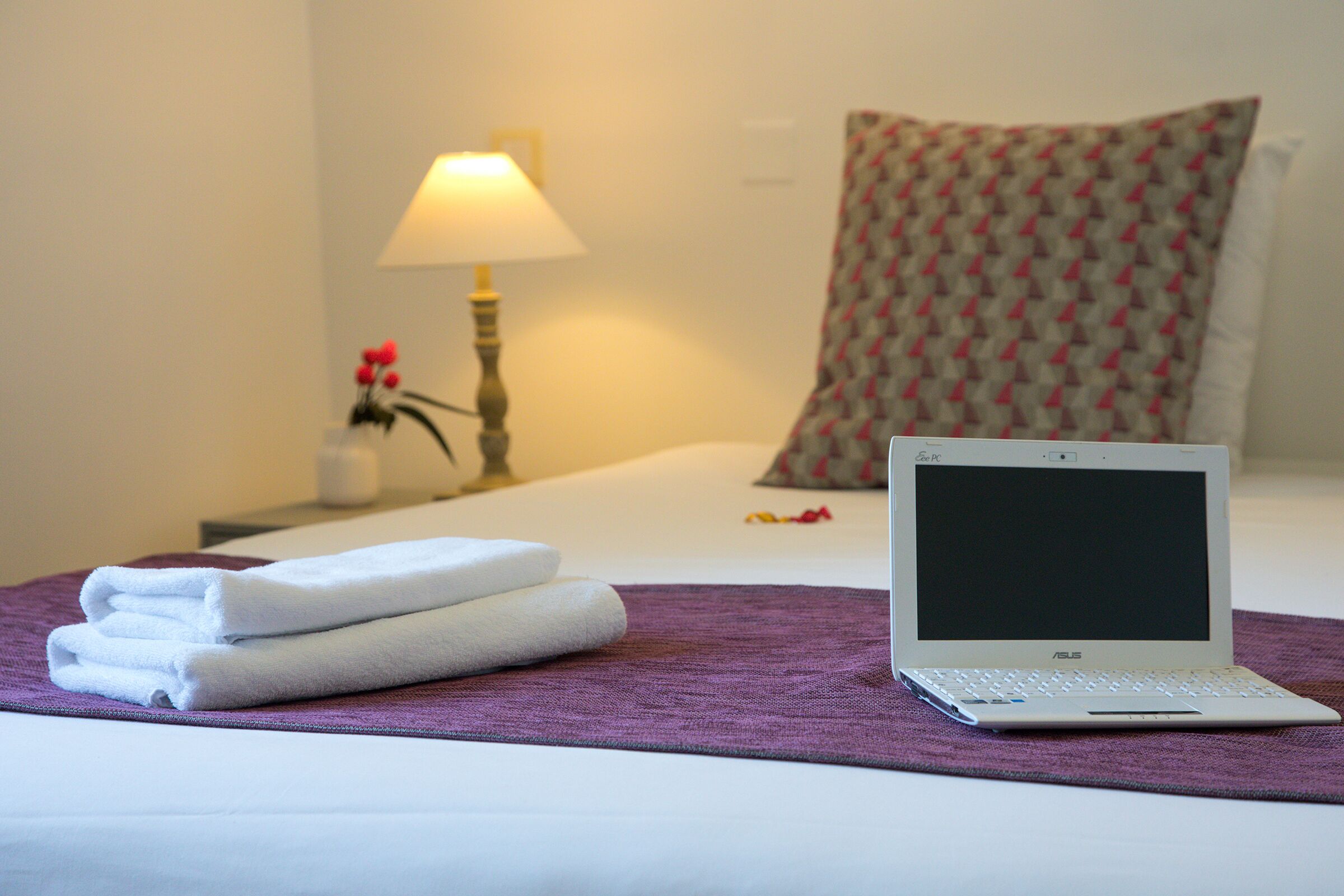 Kannettava tietokone sängyllä, jonka päällä on violetti peitto, ja sängyn vieressä on lamppu ja tyyny hotellihuoneessa.
