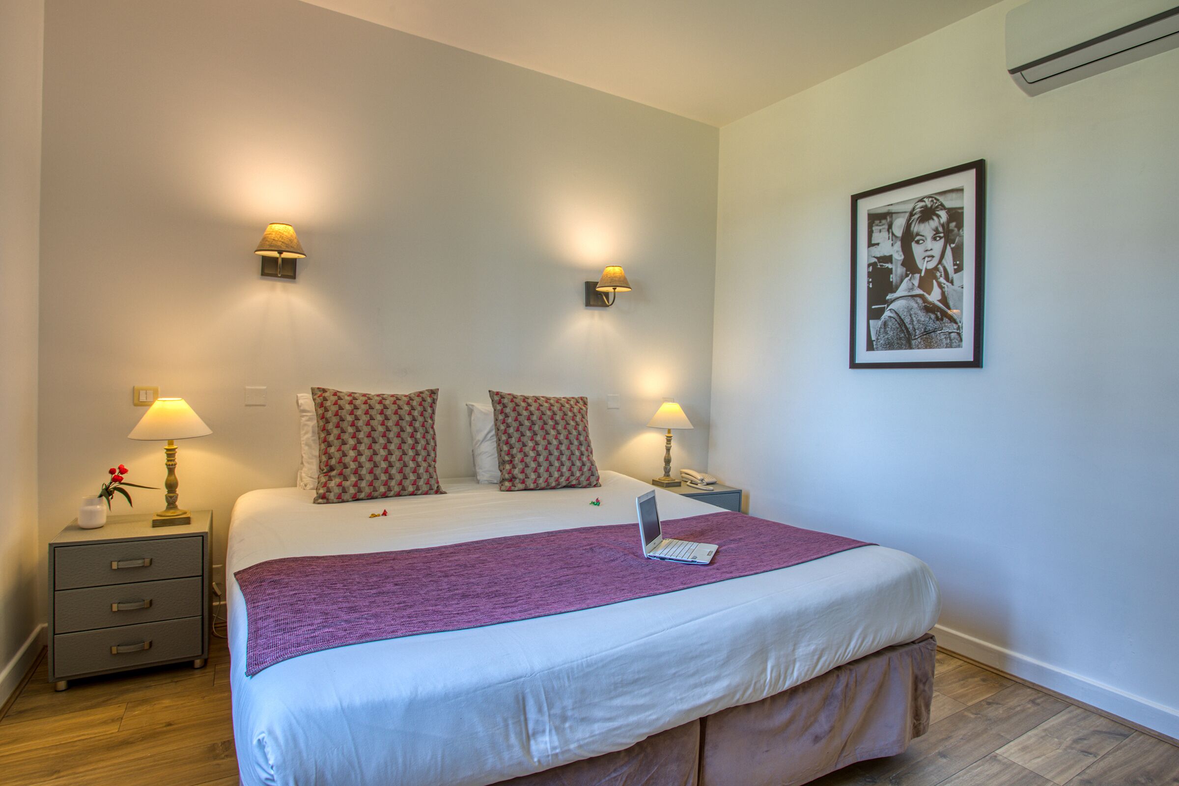 Een netjes opgemaakt bed met een laptop, twee bedlampjes, een ingelijst foto aan de muur, en een klein nachtkastje met een lamp en een vaas.