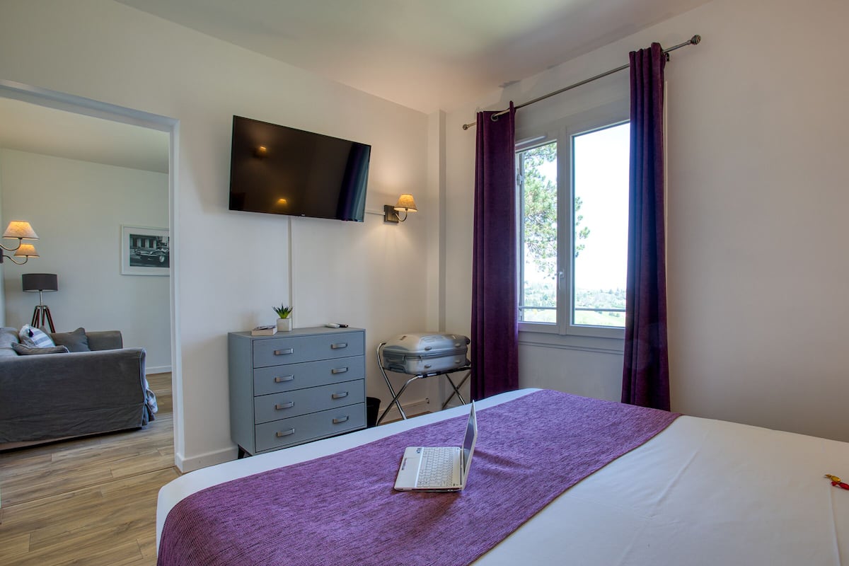 Une chambre à coucher avec un lit, un ordinateur portable, une commode, un téléviseur et une fenêtre avec des rideaux.