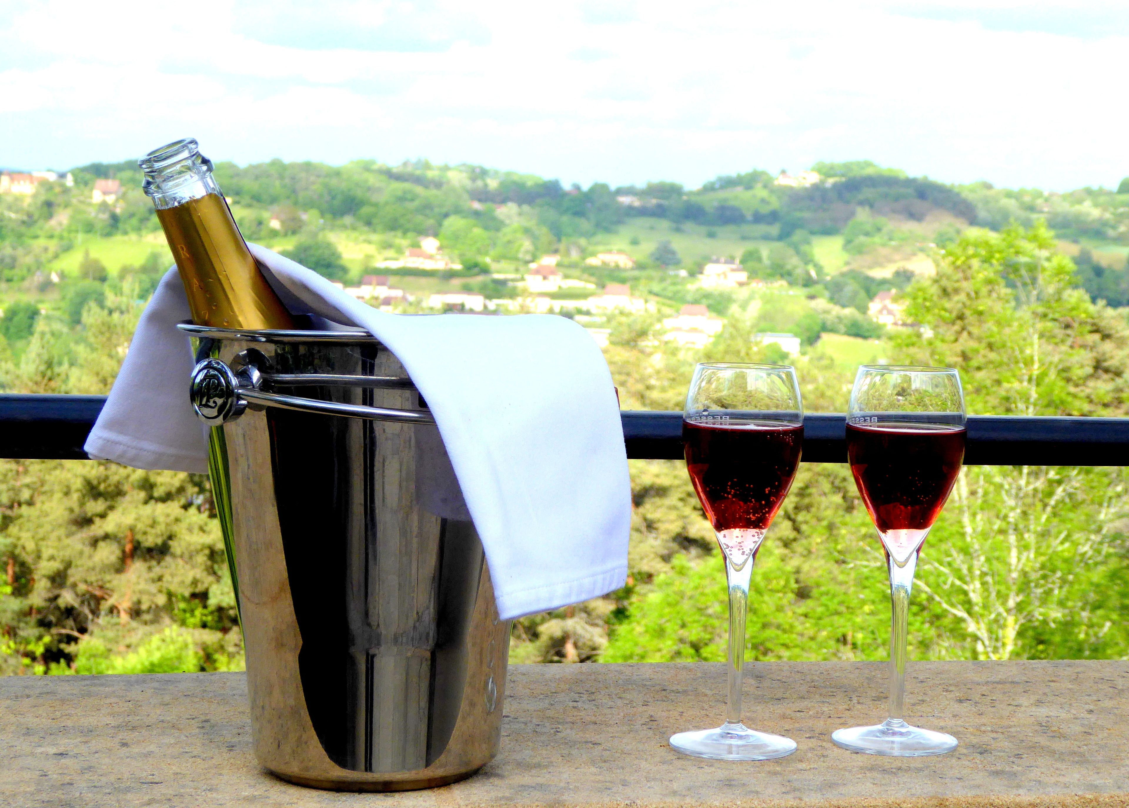 A bottle of champagne in a metal bucket with a white cloth, two wine glasses filled with red wine, and a scenic view of a green landscape with houses in the background.