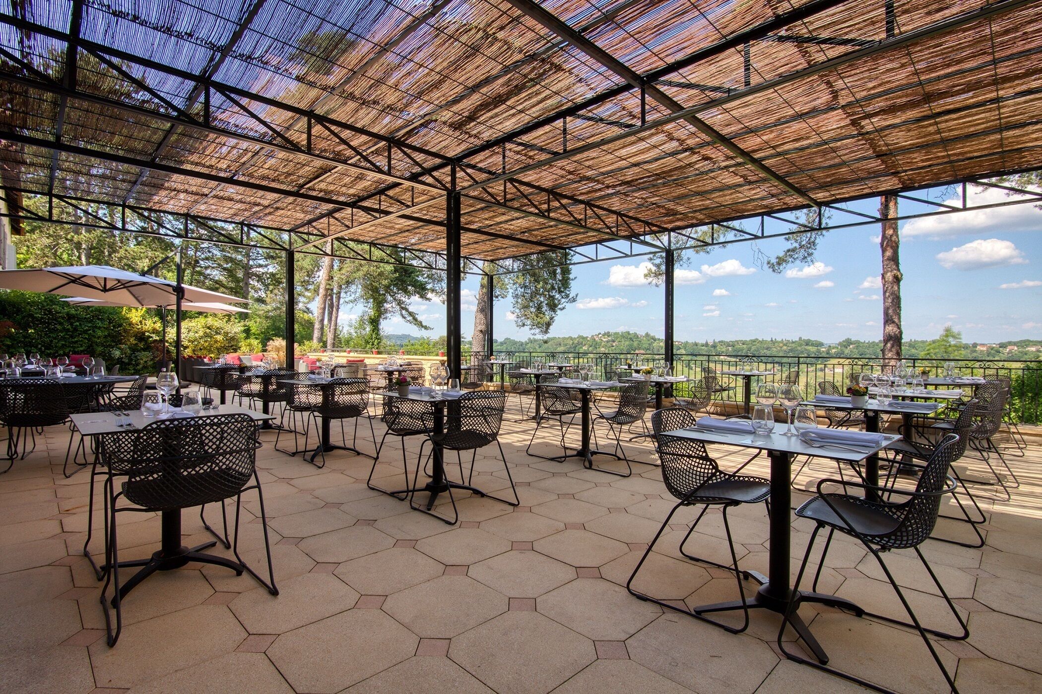 Un patio exterior con baldosas hexagonales, mesas y sillas de metal, y vistas a un paisaje natural.