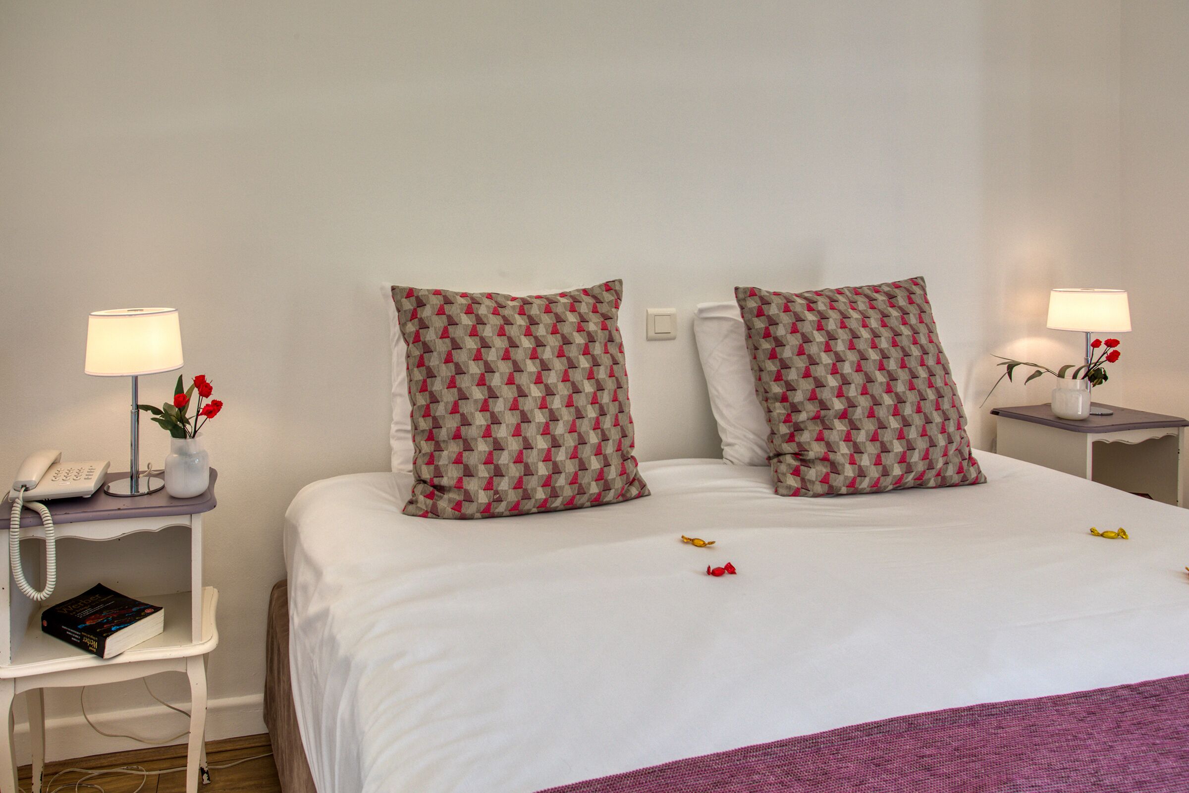 A neatly made bed with patterned pillows, a bedside table with a lamp, a phone, and a vase with flowers.