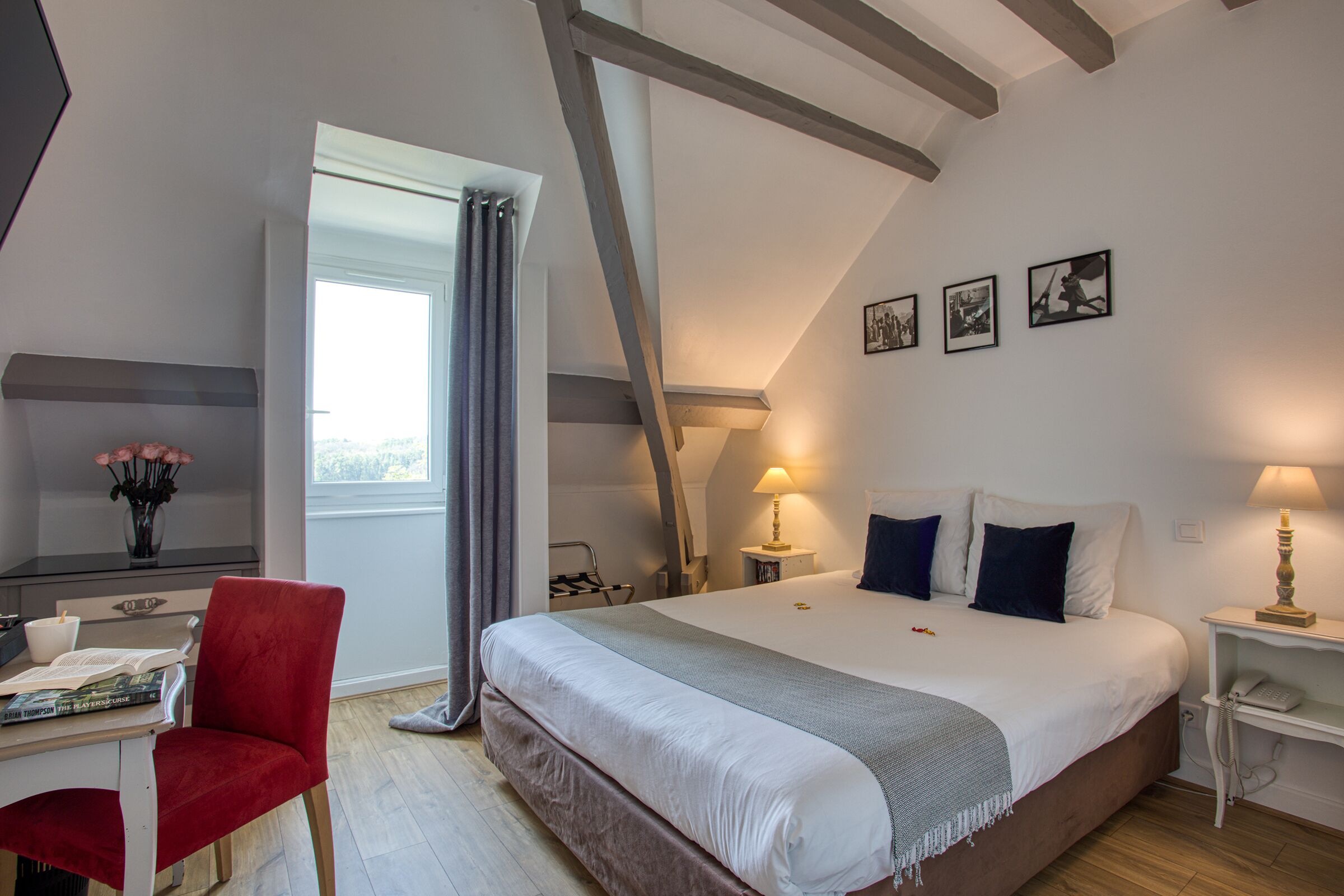 A bedroom with a large bed, a desk with a chair, a window with curtains, and a lamp on a bedside table.