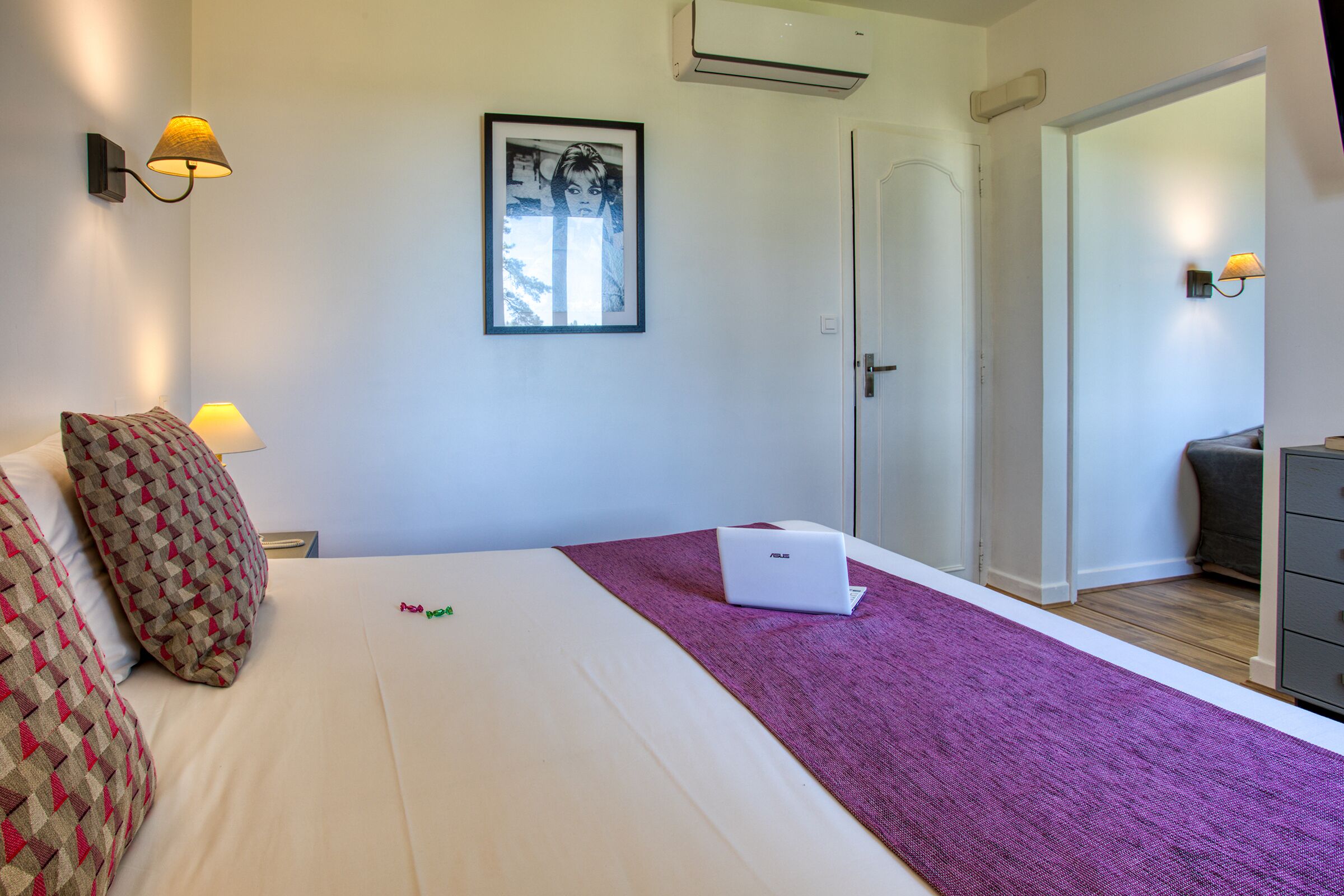 A hotel room with a bed, a laptop, a purple throw, and a framed picture on the wall.