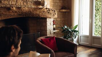 A man reading a book in a cozy room with a brick fireplace, a red cushion, and a plant by the window.