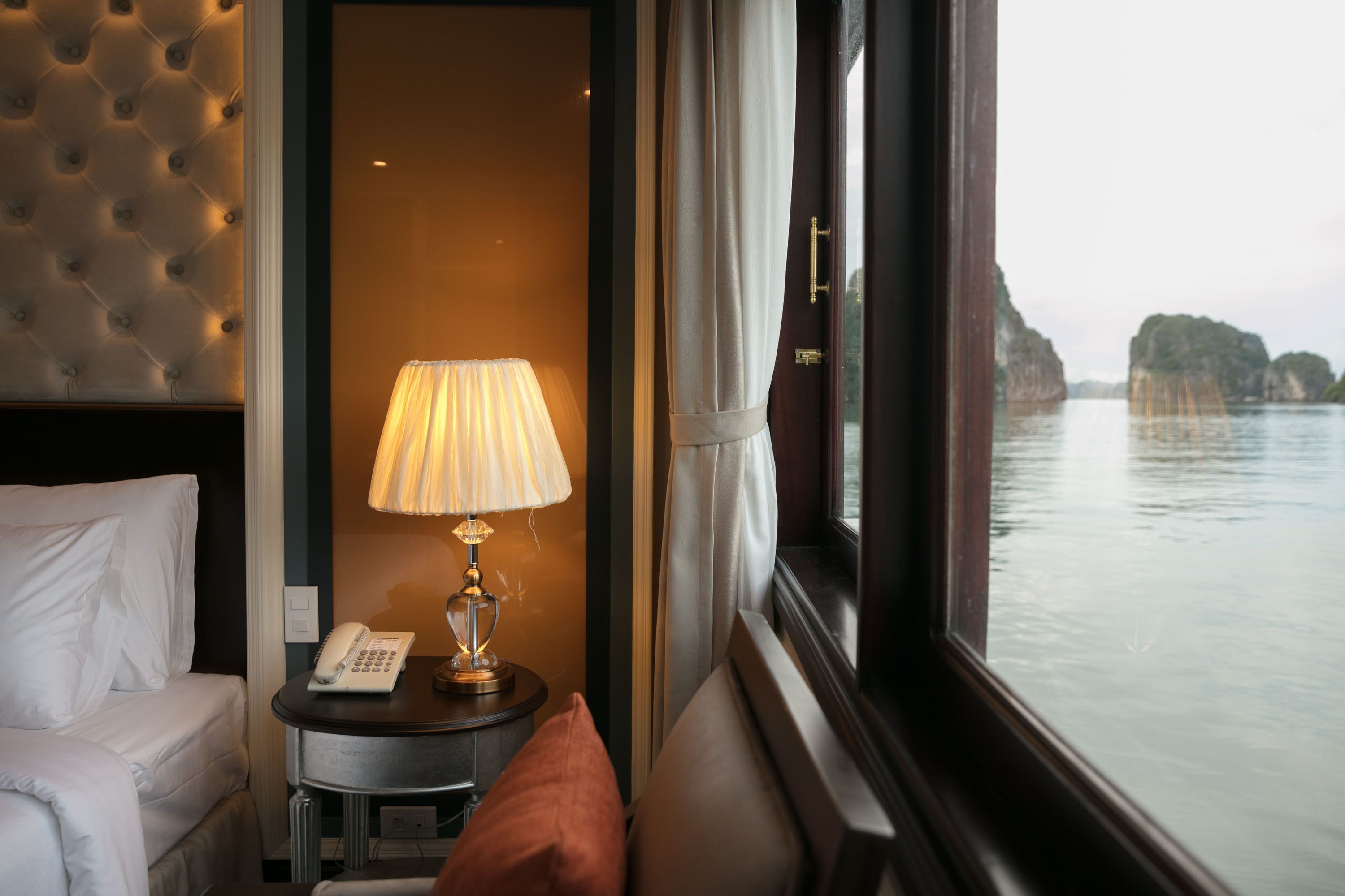 Habitación de hotel con cama, mesita de noche con lámpara, teléfono y vista a un cuerpo de agua a través de una ventana.