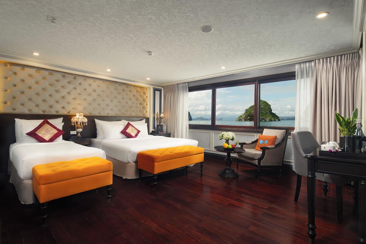 A hotel room with two beds, a sitting area with a chair and table, and a large window with a view of the sea.