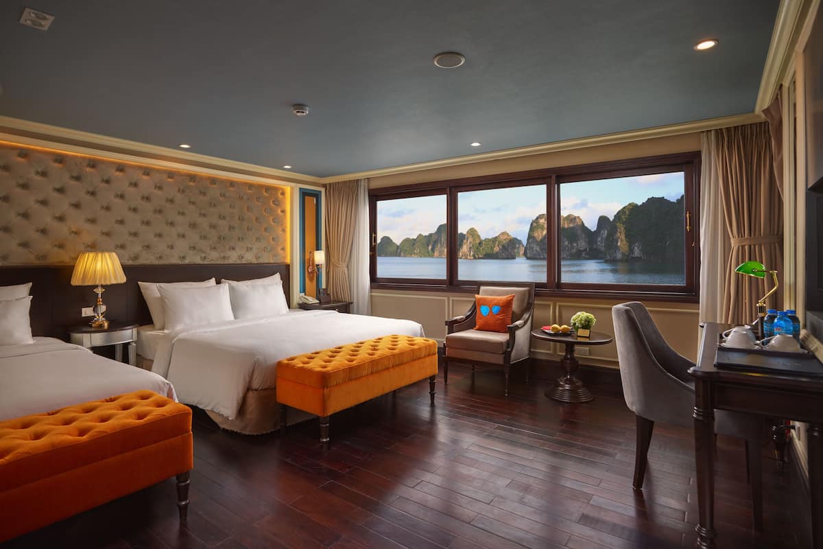 A hotel room with two beds, a large window with a view of a bay, a desk with a chair, and a small table with a lamp.