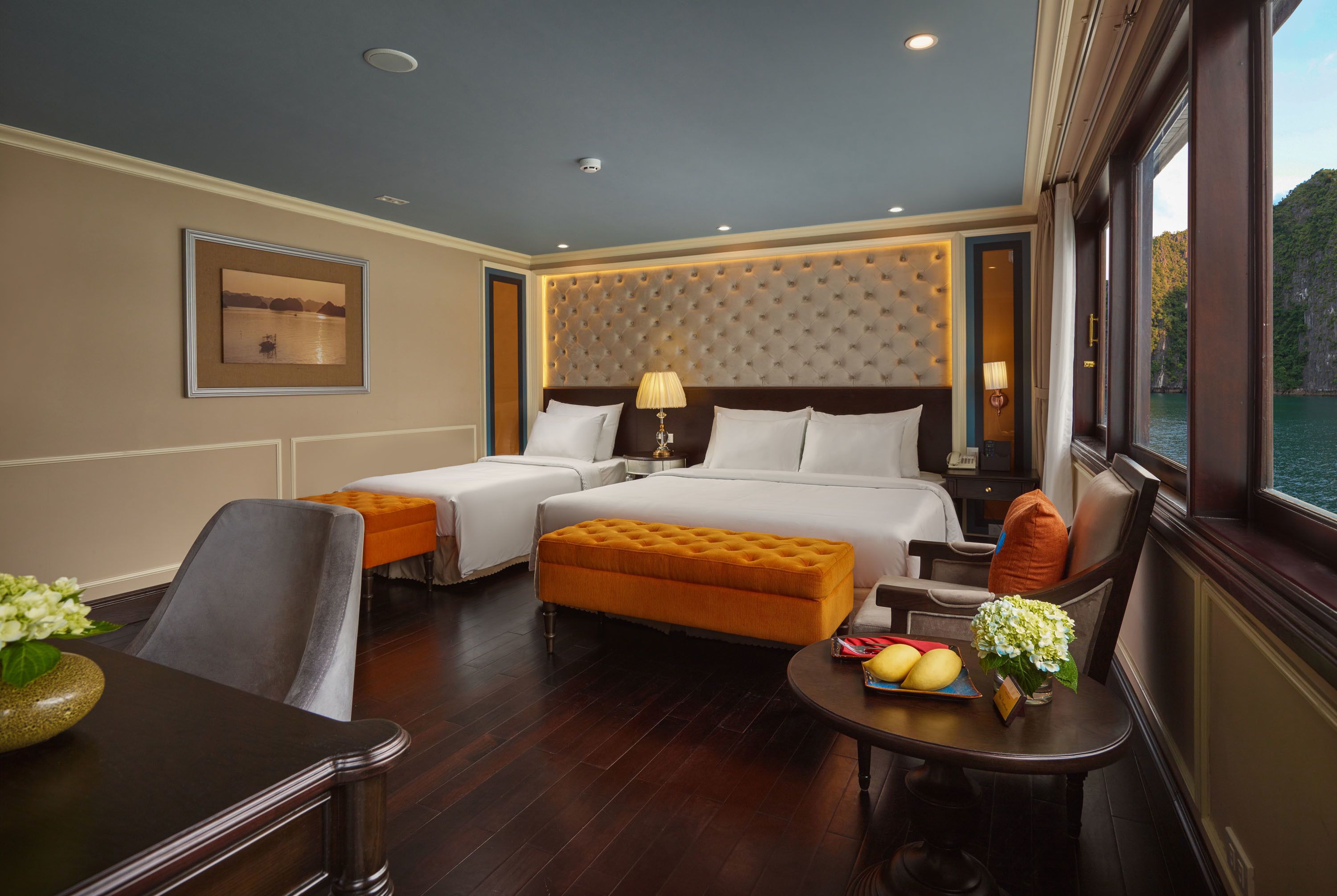 Quarto de hotel com uma cama grande, duas mesas de cabeceira, um sofá, uma mesa de jantar com frutas e flores, e vista para a água.