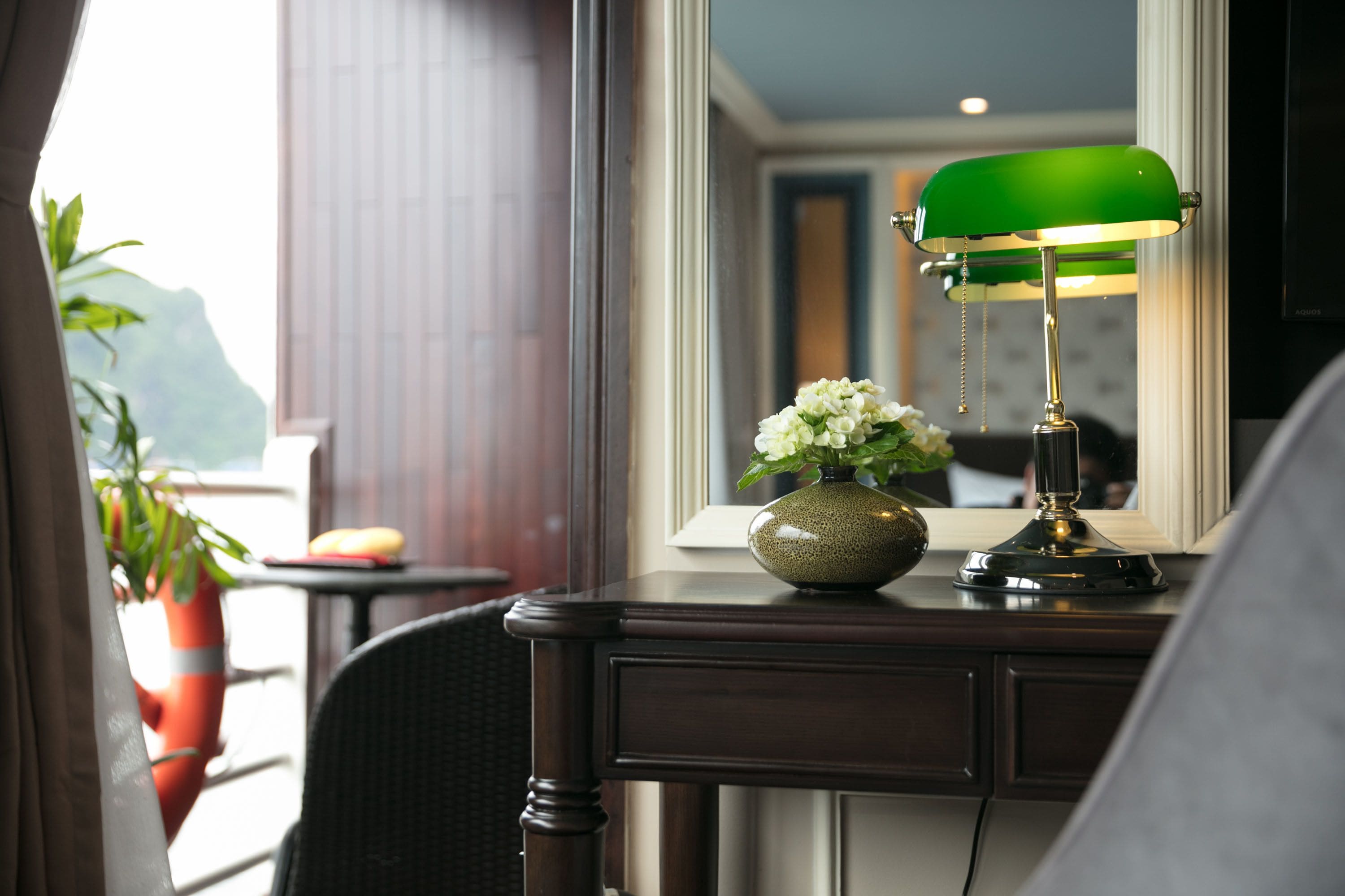 A hotel room with a desk, a green desk lamp, a vase with flowers, and a view of the outdoors.