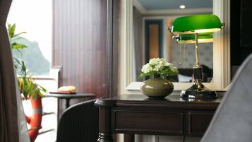 A hotel room with a desk, a green desk lamp, a vase with flowers, and a view of the outdoors.