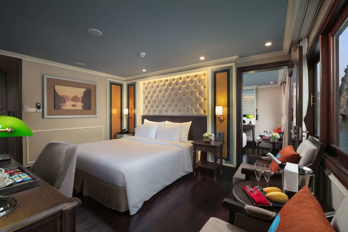 A hotel room with a large bed, a desk with a lamp, a dining area with a table and chairs, and a view of the outdoors.