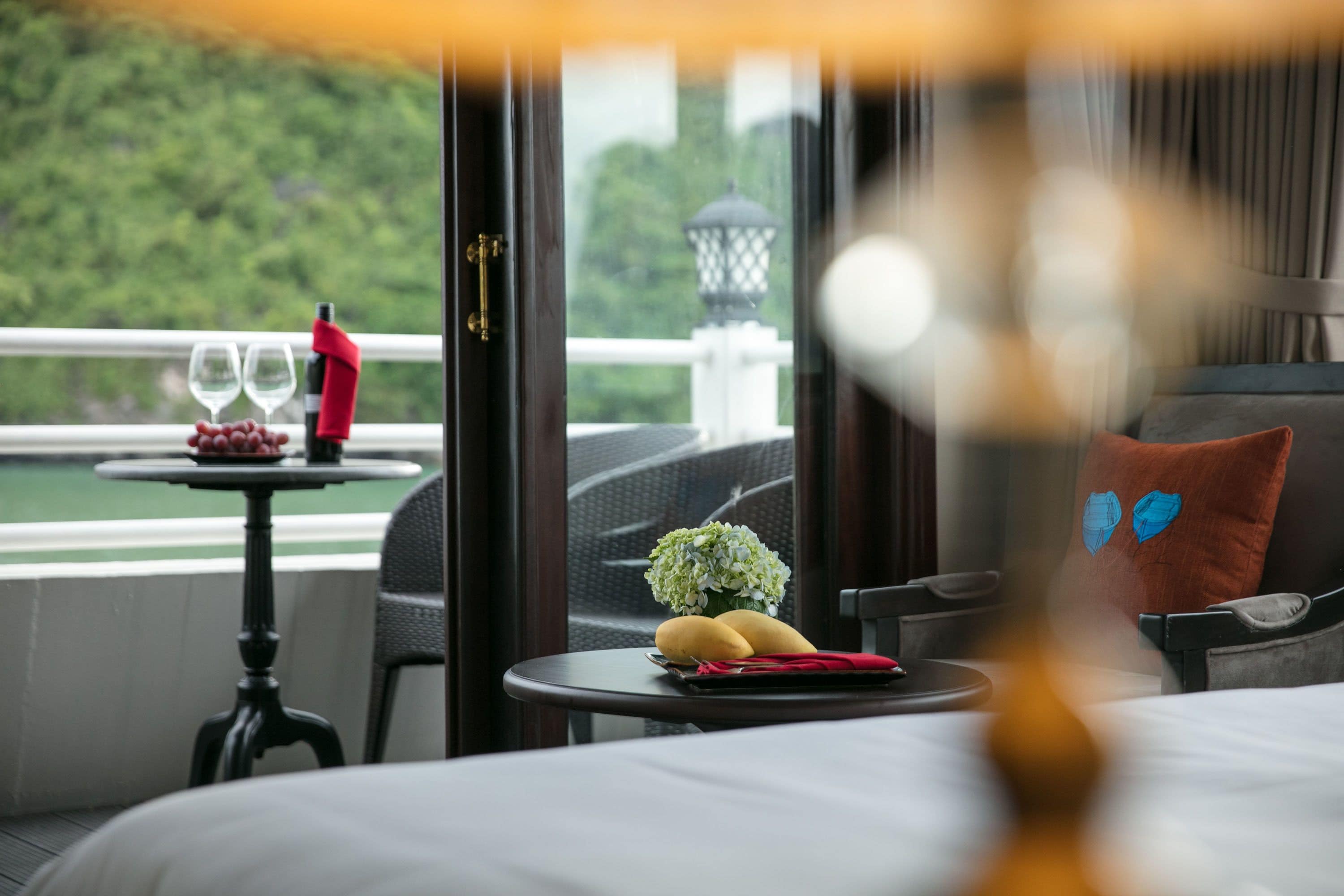 A hotel room with a bed, a small table with a bottle and glasses, and a view of greenery outside.