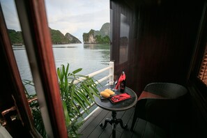 A balcony with a table set for two, a plant, and a view of the water and mountains. - Halong Athena Cruise (Hon Gai)