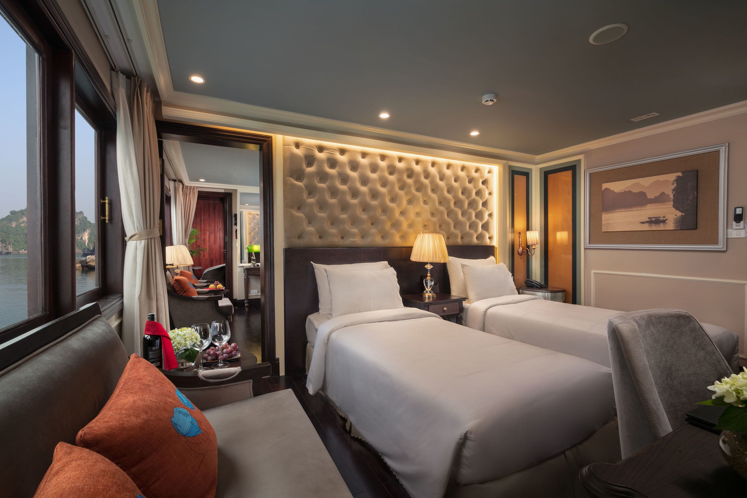 A hotel room with two beds, a large window with curtains, a view of the water, a headboard with a textured pattern, and a painting on the wall.
