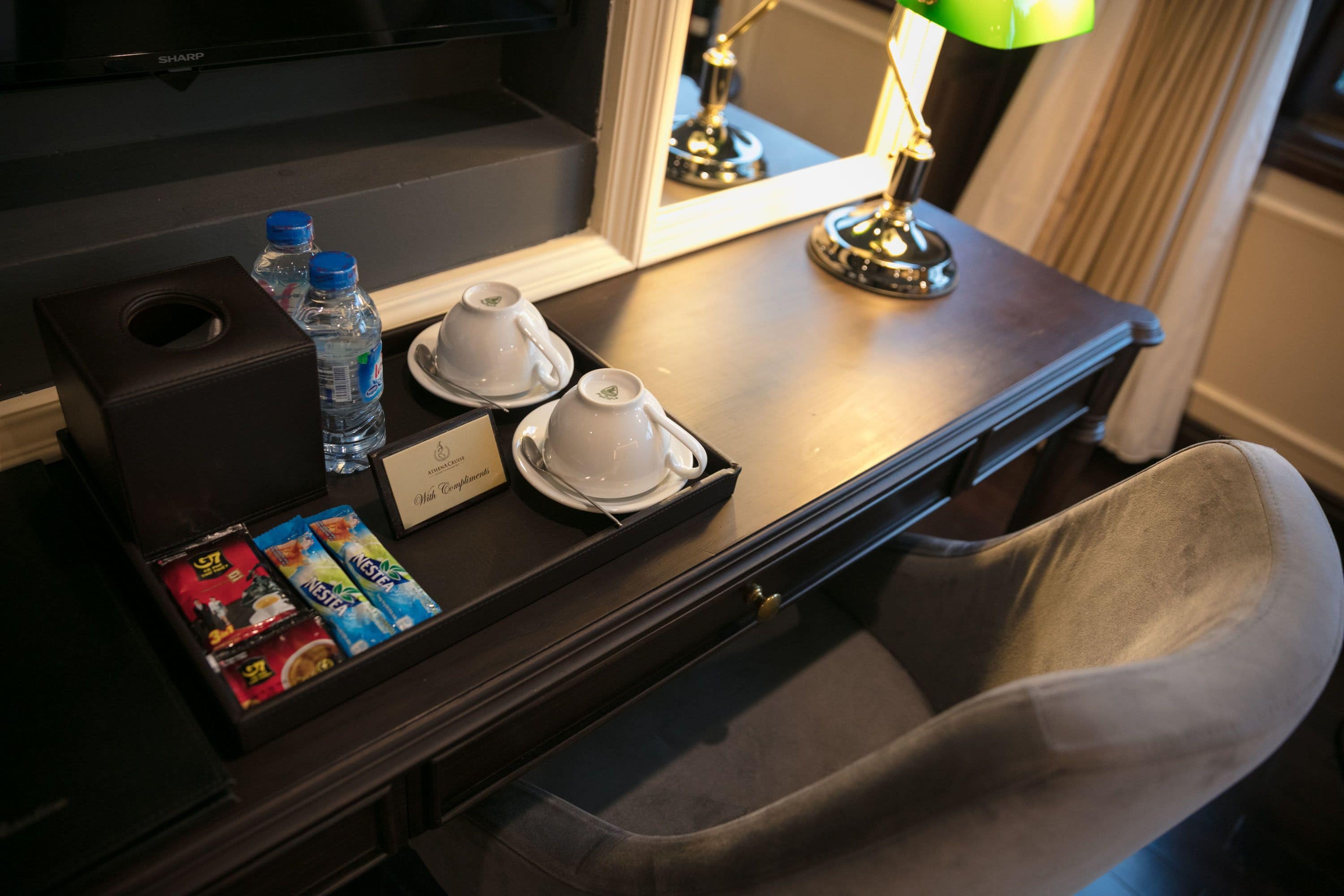 A hotel room desk with a tray containing two white cups, two water bottles, a box of tissues, and a tray of assorted chocolates.