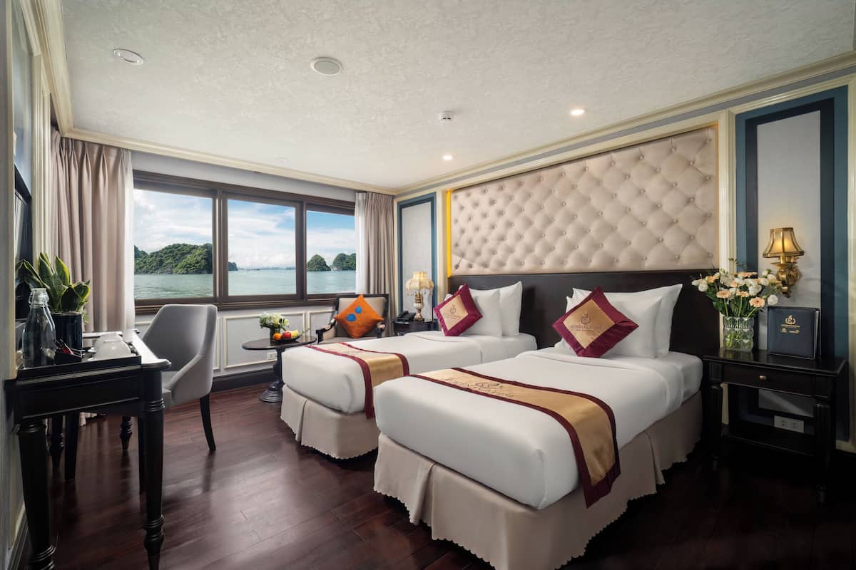 A hotel room with two beds, a desk, a chair, and a view of the sea.