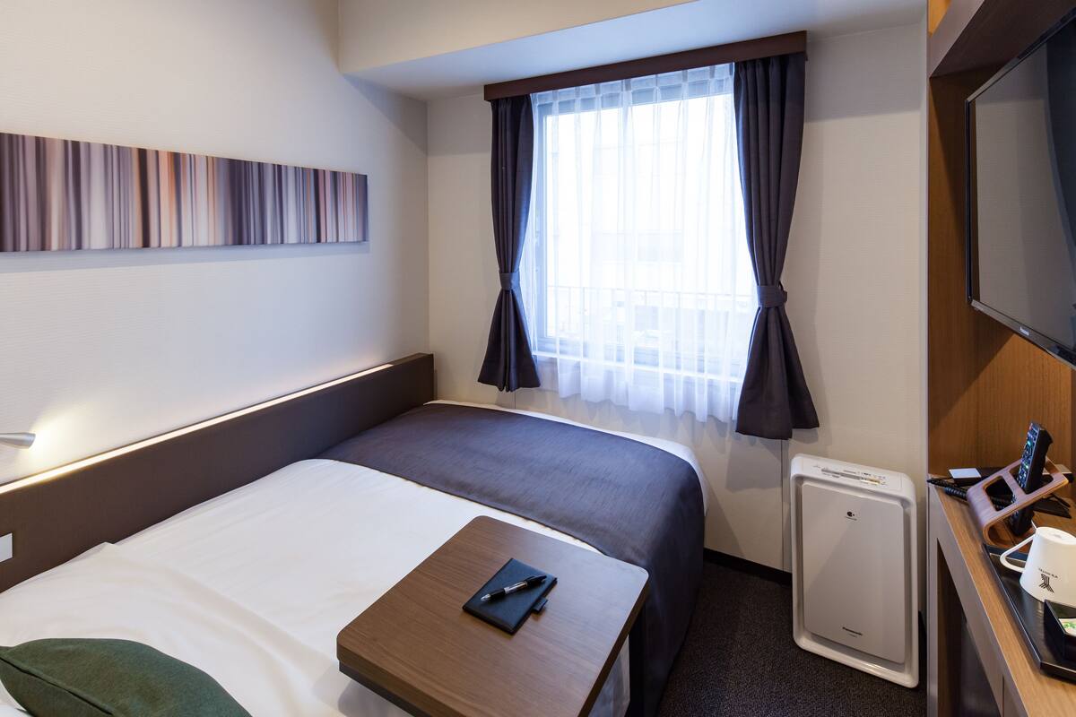 A hotel room with a bed, a desk with a computer, a small refrigerator, and a window with curtains.