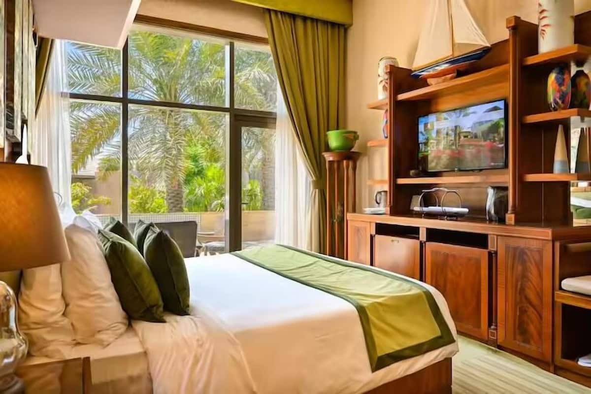 A hotel room with a bed, a television on a wooden cabinet, and a view of palm trees through large windows.