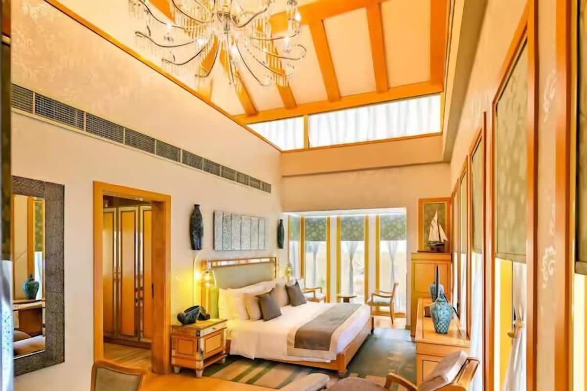 A spacious bedroom with a large bed, bedside tables, a desk, and a chair. There is a chandelier on the ceiling and large windows providing natural light.