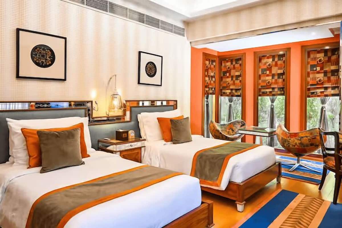 A hotel room with two beds, a small table, a chair, and a window with a view of trees.