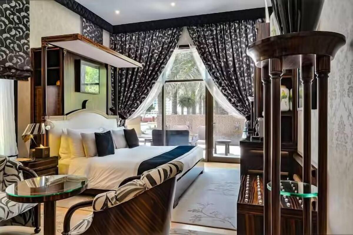 A hotel room with a large bed, a glass table, a chair, a desk, and a view of the outdoors.