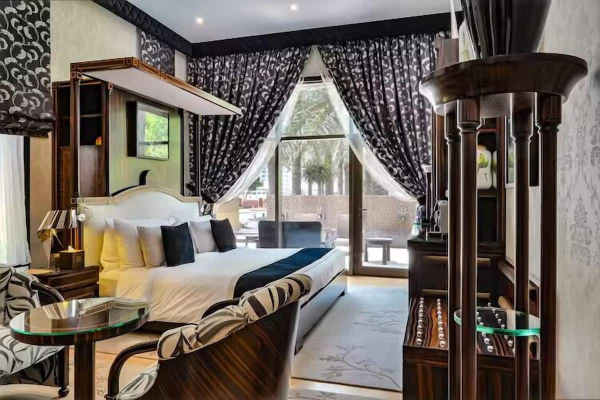 A hotel room with a bed, a glass table, a chair, a large window with curtains, and a view of the outside.