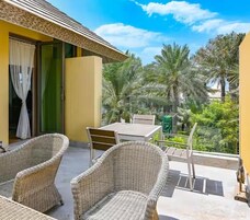 Un balcón con mobiliario de mimbre, una mesa y sillas, con vista a un jardín con palmeras.