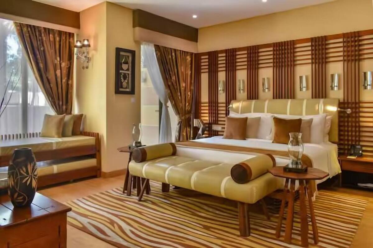 A spacious bedroom with a large bed, a sofa, and a bench. There is a window with curtains, a wall-mounted light fixture, and a decorative vase on a table.