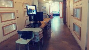 A long, narrow hallway with a series of desks and chairs, each equipped with a computer monitor and keyboard. The hallway is lined with doors on both sides, some with blinds. - Life Guesthouse - Hostel (Tongyeong)