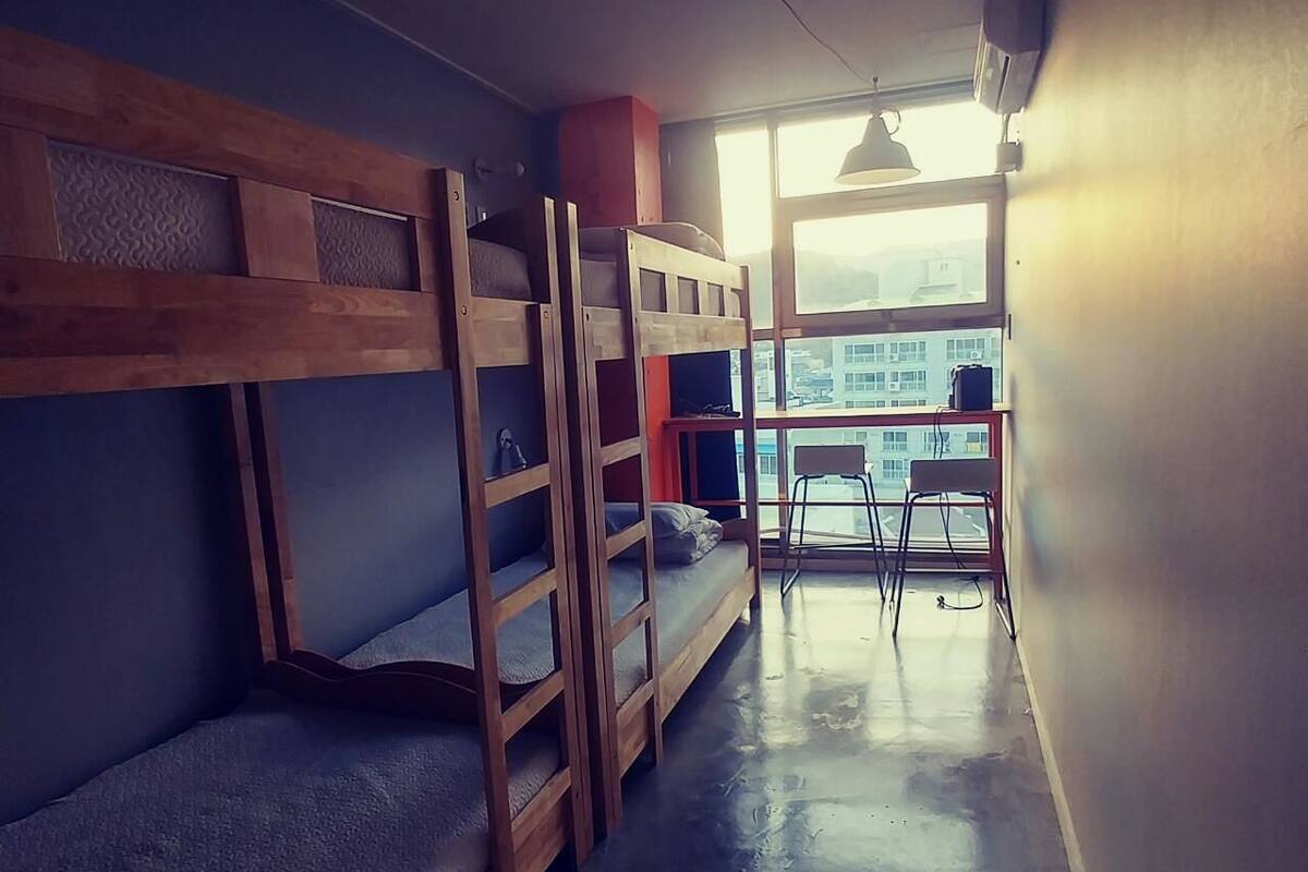 A dormitory room with bunk beds, a small table, and a chair near a window.