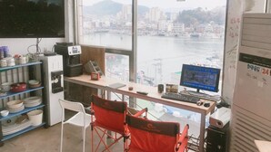 A room with a large window overlooking a cityscape and water. There is a desk with a computer, a chair, and shelves with dishes. - Life Guesthouse - Hostel (Tongyeong)