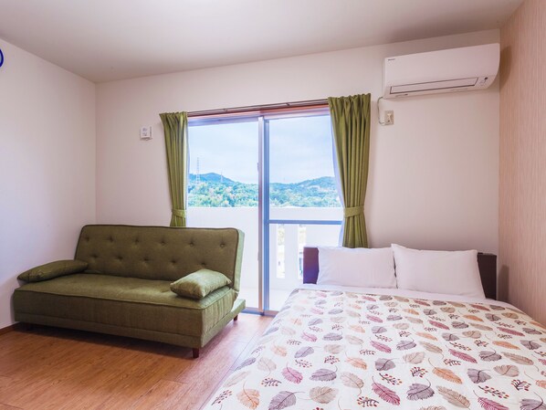 A room with a bed, a green sofa, and a view of a mountainous landscape through a large window. - Kariyushi Condominium Resort Kin Yaka Seaside (Kin)